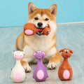 Shiba Inu dog holding an orange latex squeaky toy in its mouth, with three more cute latex animal chew toys (white cow, purple hippo, brown dog) lined up in front.