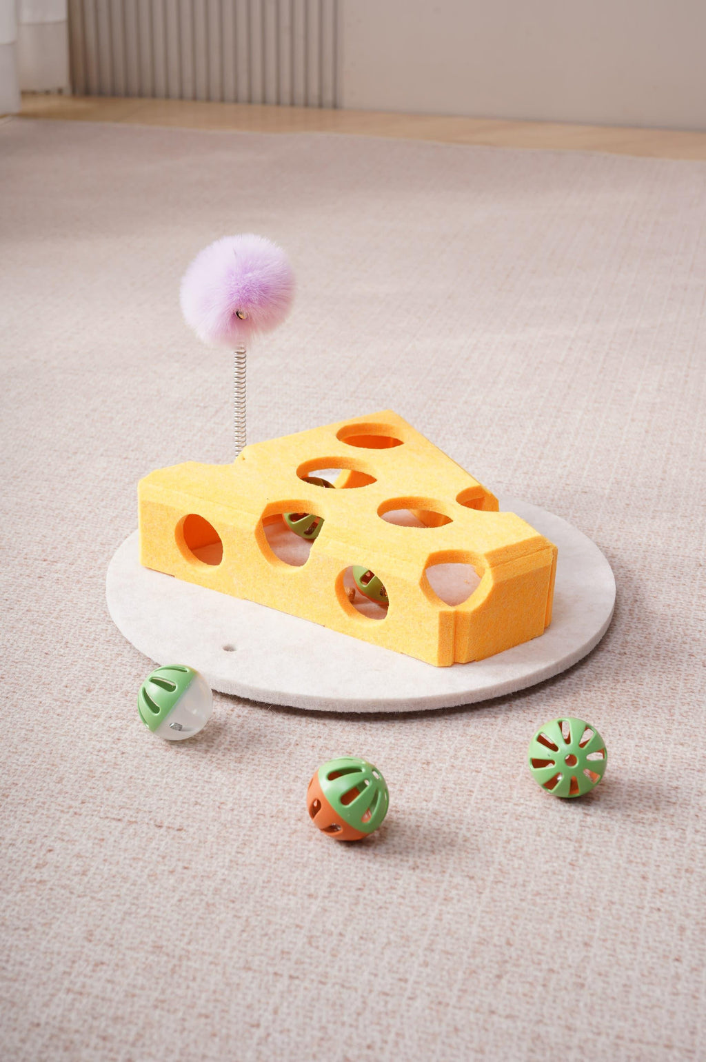 Yellow cheese-shaped felt cat toy with holes, purple fuzzy ball on a spring wand, and three small balls on a beige carpet.