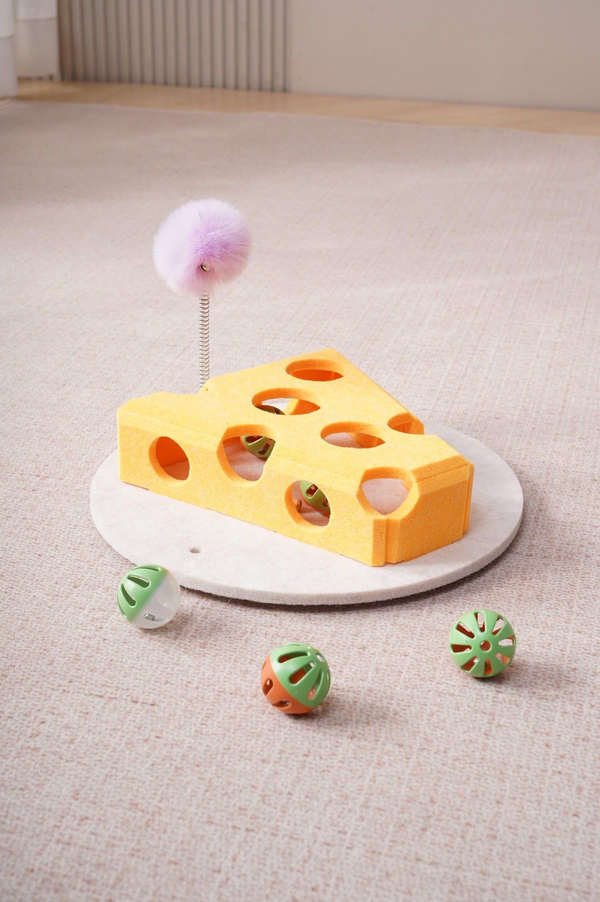 Yellow cheese-shaped felt cat toy with holes, purple fuzzy ball on a spring wand, and three small balls on a beige carpet.