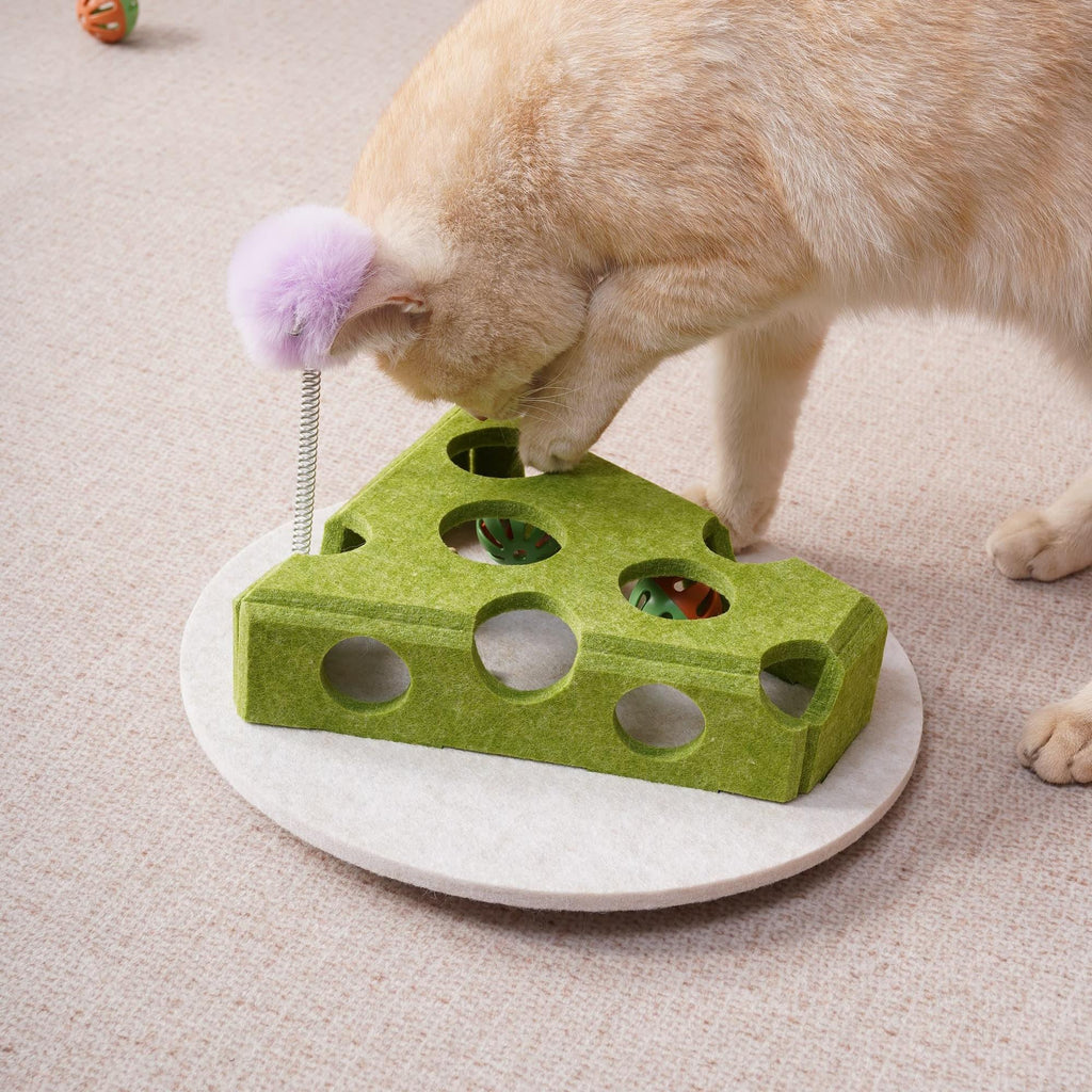 Cream-colored cat leaning over the green cheese-shaped felt cat toy, pawing at balls through its holes, with a purple fuzzy ball on a spring wand nearby.