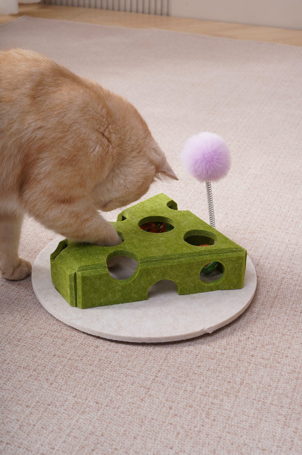 Orange cat batting balls through the holes of the green cheese-shaped felt cat toy with a purple fuzzy ball on a spring wand, on beige carpet.