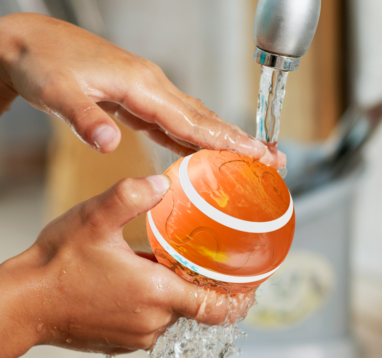 Hands washing an orange intelligent self-rotating cat ball with white stripes under running faucet water, showing waterproof design.