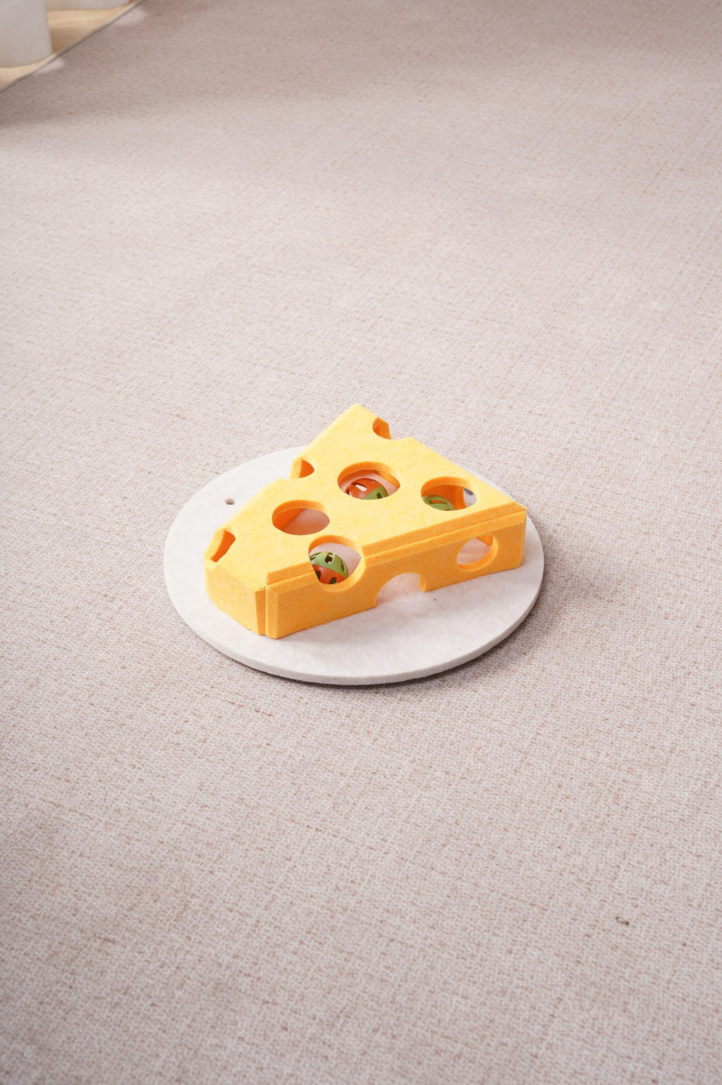 Overhead view of the yellow cheese-shaped felt cat toy with colorful balls visible through its holes, on a round base over beige carpet.