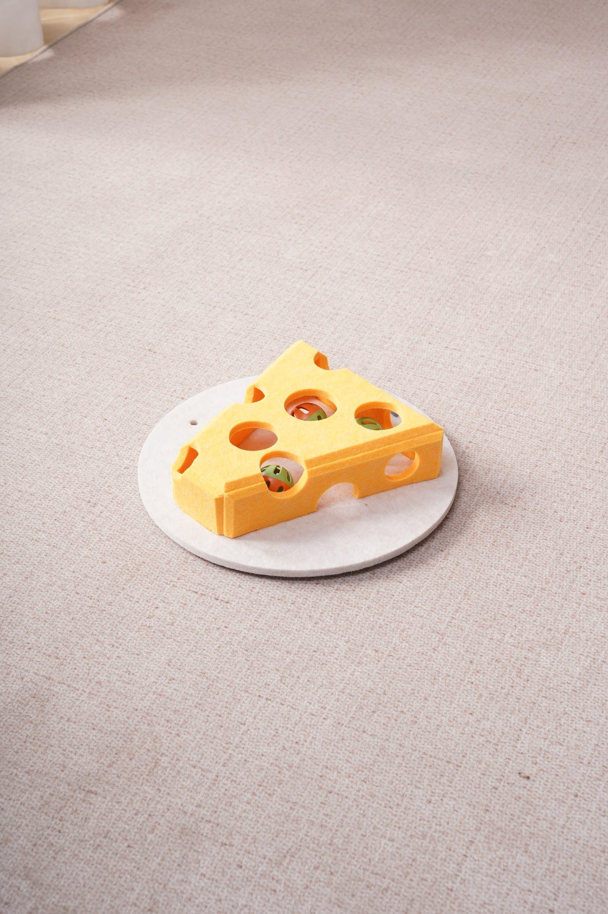 Overhead view of the yellow cheese-shaped felt cat toy with colorful balls visible through its holes, on a round base over beige carpet.