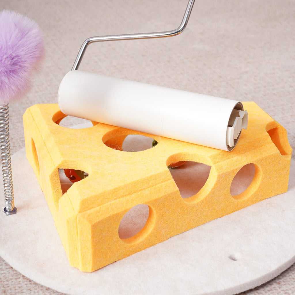 Yellow cheese-shaped felt cat puzzle toy with paper-roll tunnel on top and purple fuzzy ball on a spring wand, on a round base.
